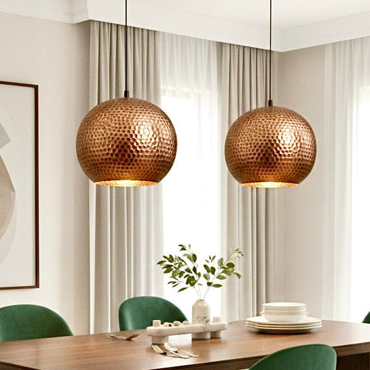 Two handmade hammered copper pendant lights over a modern wooden dining table with green velvet chairs.