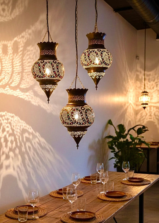 Trio of 100% handmade Moroccan brass lamps hanging over a rustic wooden dining table, casting intricate geometric shadows on the wall.