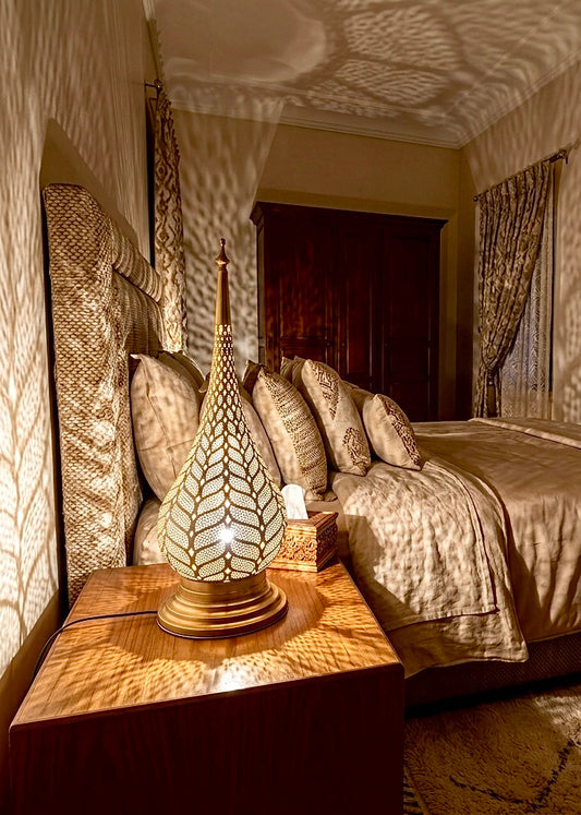 An ornate Moroccan brass bedside lamp on a nightstand, casting decorative shadows across a cozy, traditional bedroom interior.