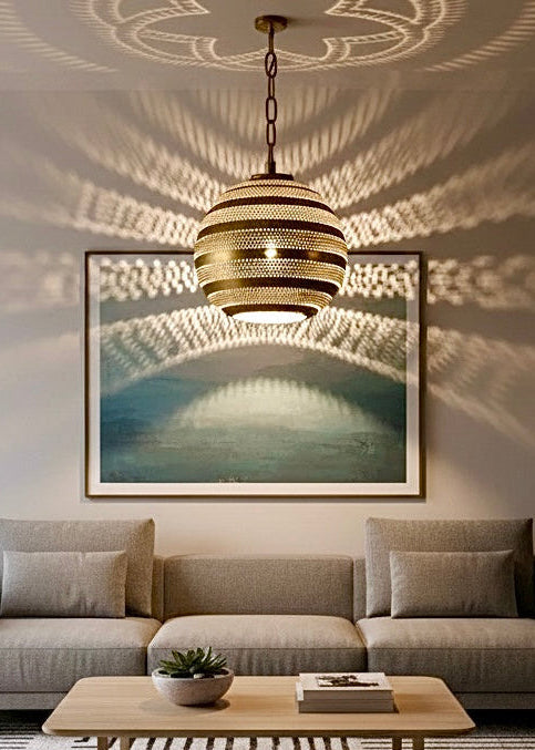 An oriental handmade brass pendant lamp casting geometric shadows in a minimalist living room with a grey sofa and wooden table.