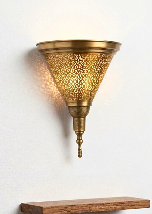 Close-up of a solid brass filigree wall sconce with warm light glowing through artisanal perforations, mounted on a clean white wall.