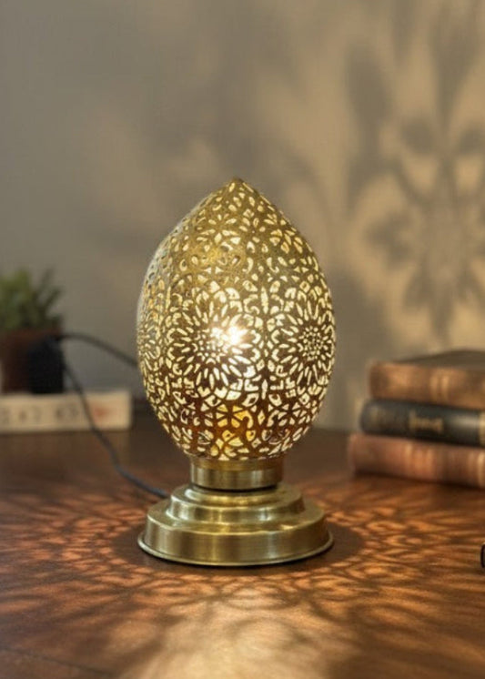 A Moroccan brass lamp glowing on a wooden vintage desk next to books, casting large floral shadows on the wall.
