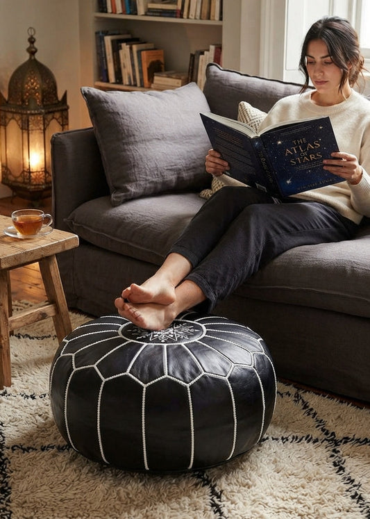 A woman resting her feet on a black Moroccan leather pouf while reading on a couch in a cozy living room.⁠