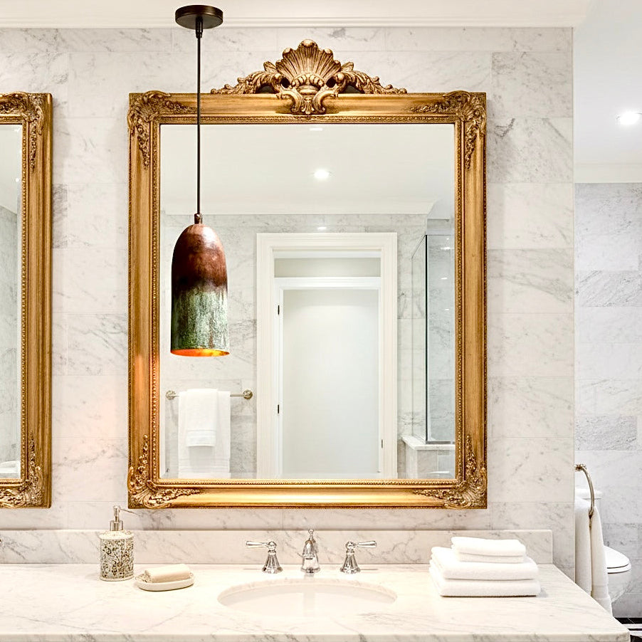 Handcrafted brass pendant fixture with green patina styled in a luxury bathroom with gold mirrors.