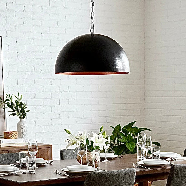 A large black hammered dome lamp with a warm copper interior lighting a dark wood dining table in a bright room.