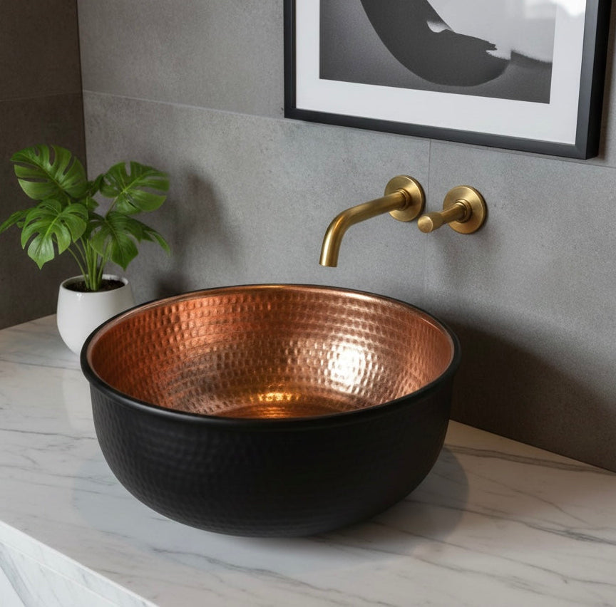 Round hammered copper vessel sink with black exterior and gold wall-mounted faucet.