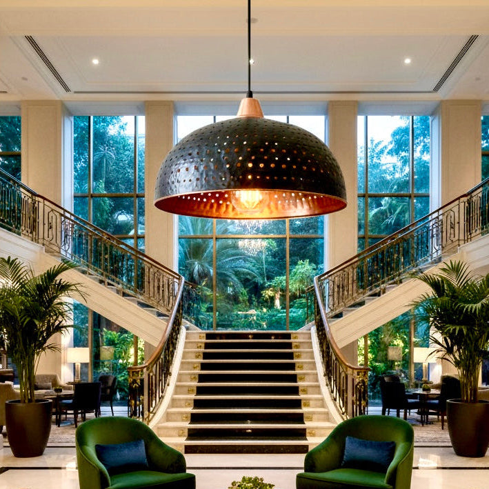 Large handmade black and brass hammered dome pendant light hanging in a luxury hotel lobby with a grand staircase.