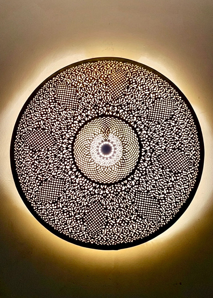 Moroccan brass wall light turned on, projecting decorative light patterns and shadows on the wall, authentic artisanal lighting for bohemian interior decor.