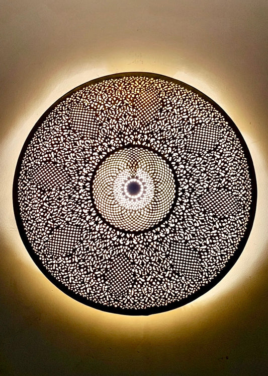 Moroccan brass wall light turned on, projecting decorative light patterns and shadows on the wall, authentic artisanal lighting for bohemian interior decor.