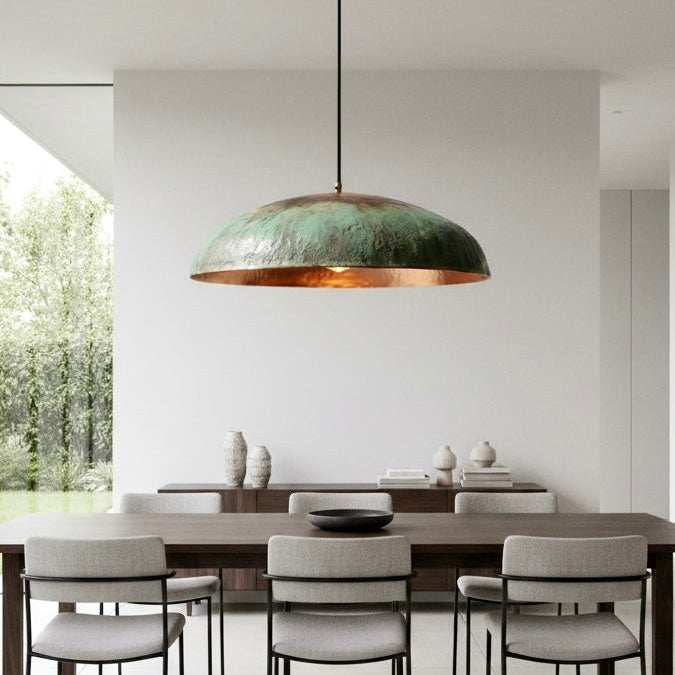 A single verdigris patina dome pendant light with a warm copper interior centered over a modern dining table in a bright room.