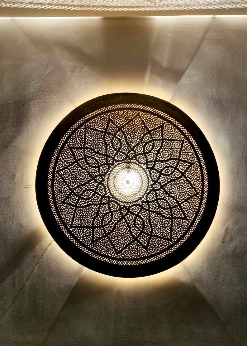 Close-up of a lit round perforated Moroccan brass lamp casting intricate geometric floral shadow patterns onto a textured wall.⁠