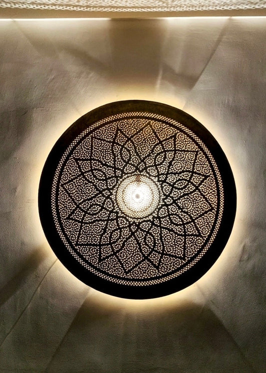 Close-up of a lit round perforated Moroccan brass lamp casting intricate geometric floral shadow patterns onto a textured wall.⁠