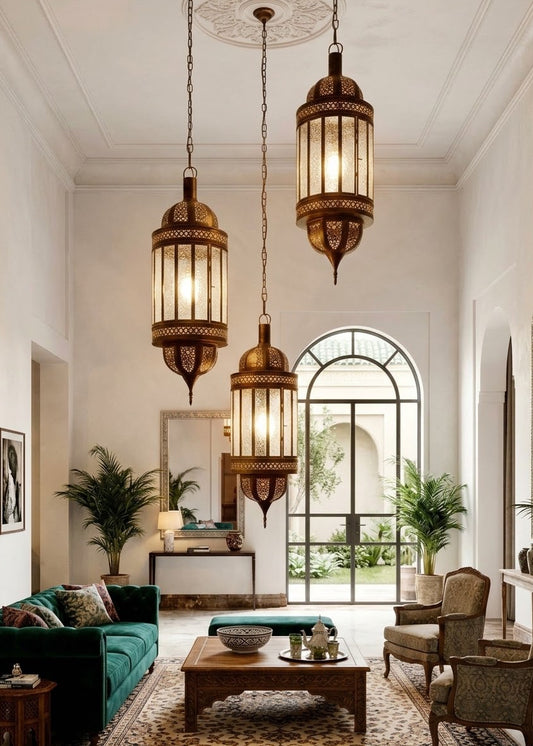 Three ornate hanging lantern pendant lights with warm glowing glass and bronze-toned brass frames, suspended in a bright, high-ceilinged living room with a green sofa, wooden coffee table, and potted plants.