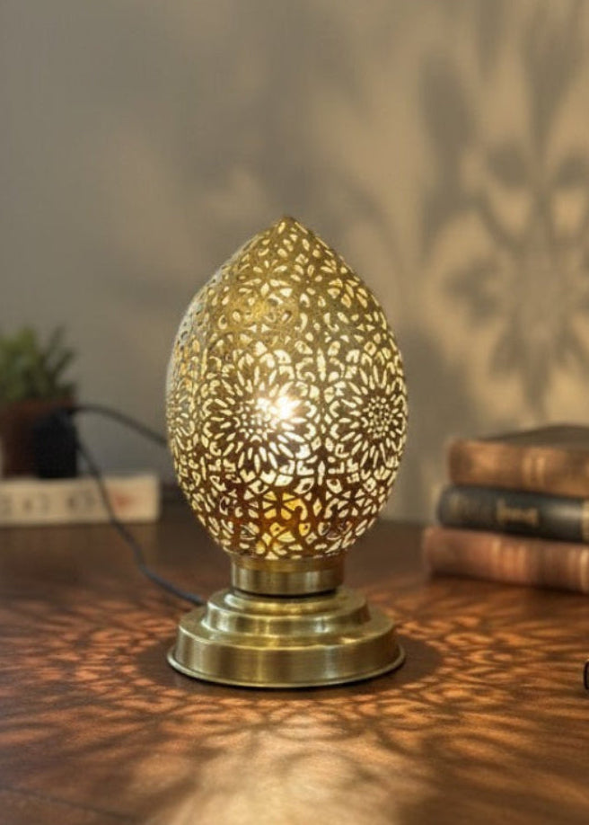 A Moroccan brass lamp glowing on a wooden vintage desk next to books, casting large floral shadows on the wall.
